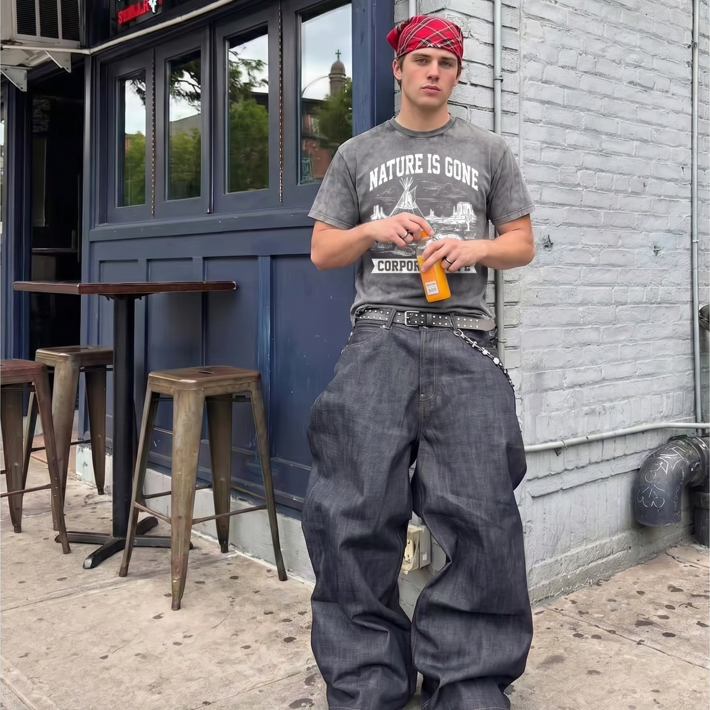 Man in oversized denim jeans, graphic tee, and red headscarf standing outside urban cafe