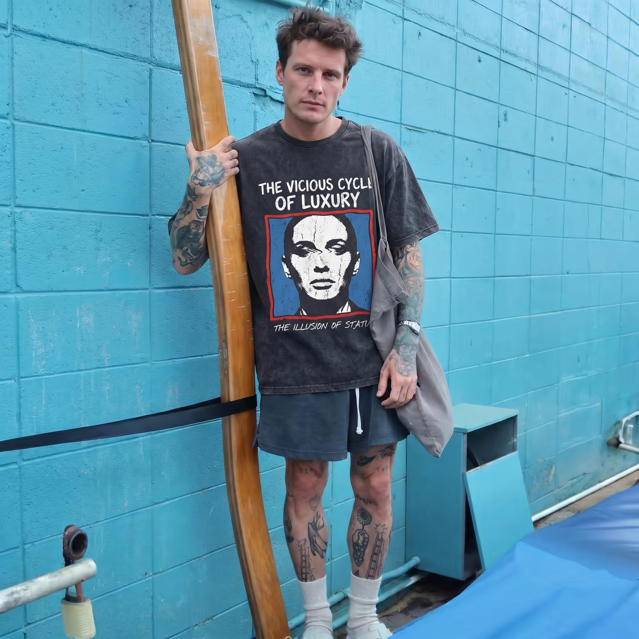 Tattooed man in graphic t-shirt, shorts, and socks holding a surfboard by a blue wall