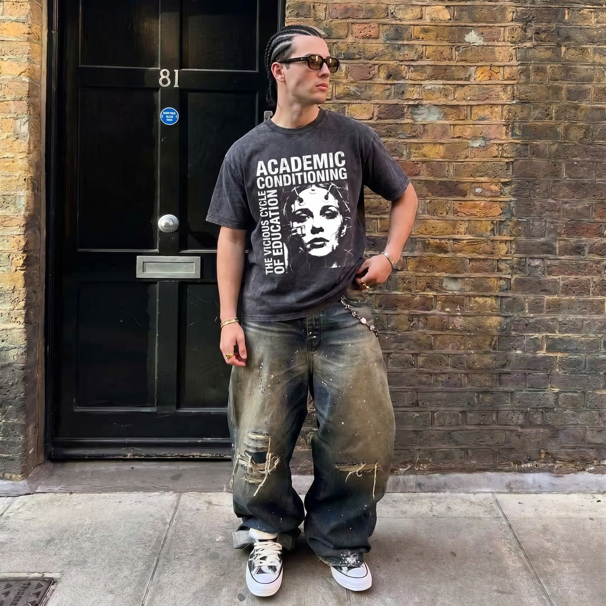 Man in graphic 'Academic Conditioning' t-shirt, baggy distressed jeans, and sneakers standing in front of a brick wall and black door.