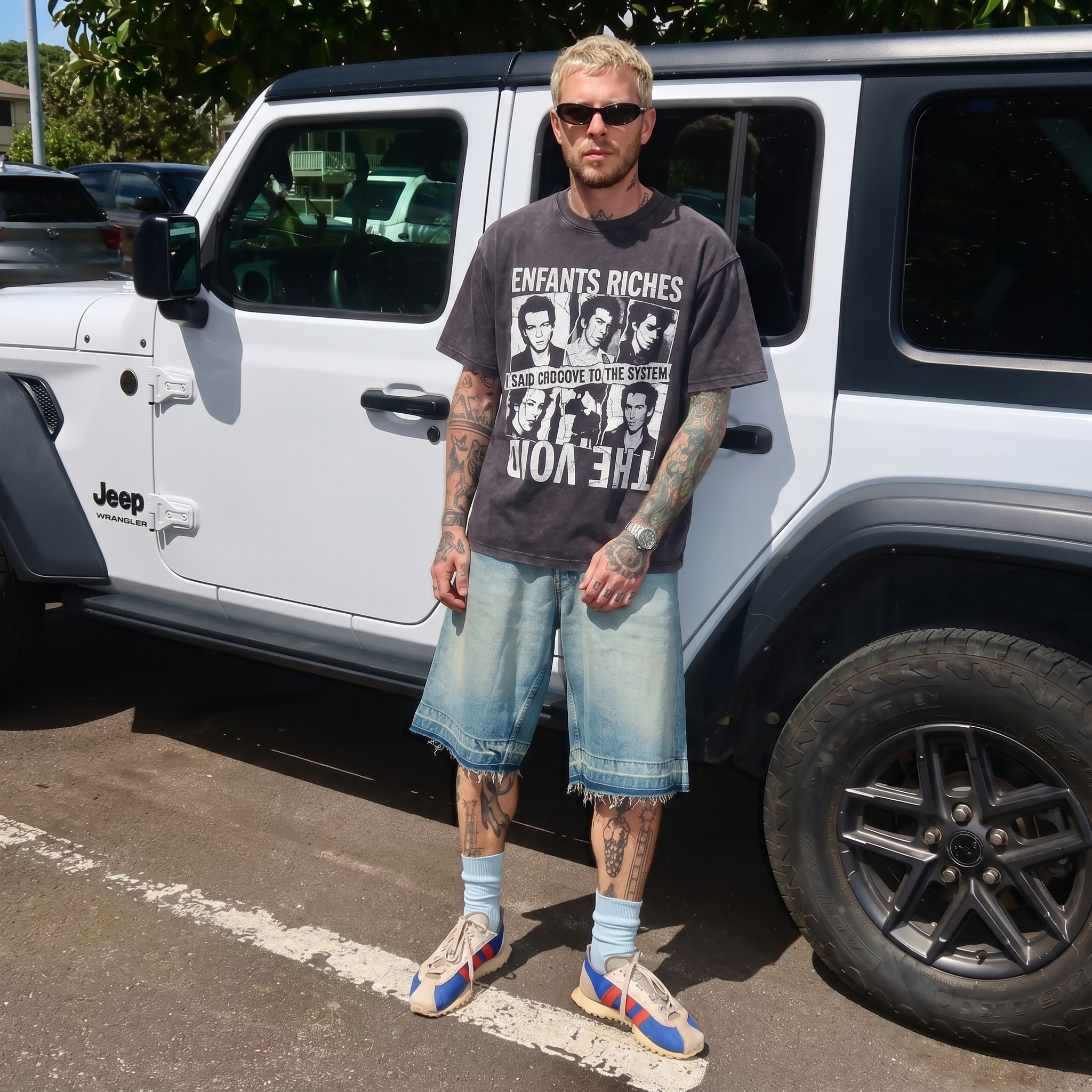 Tattooed man in graphic T-shirt, denim shorts, and sunglasses stands by white Jeep Wrangler