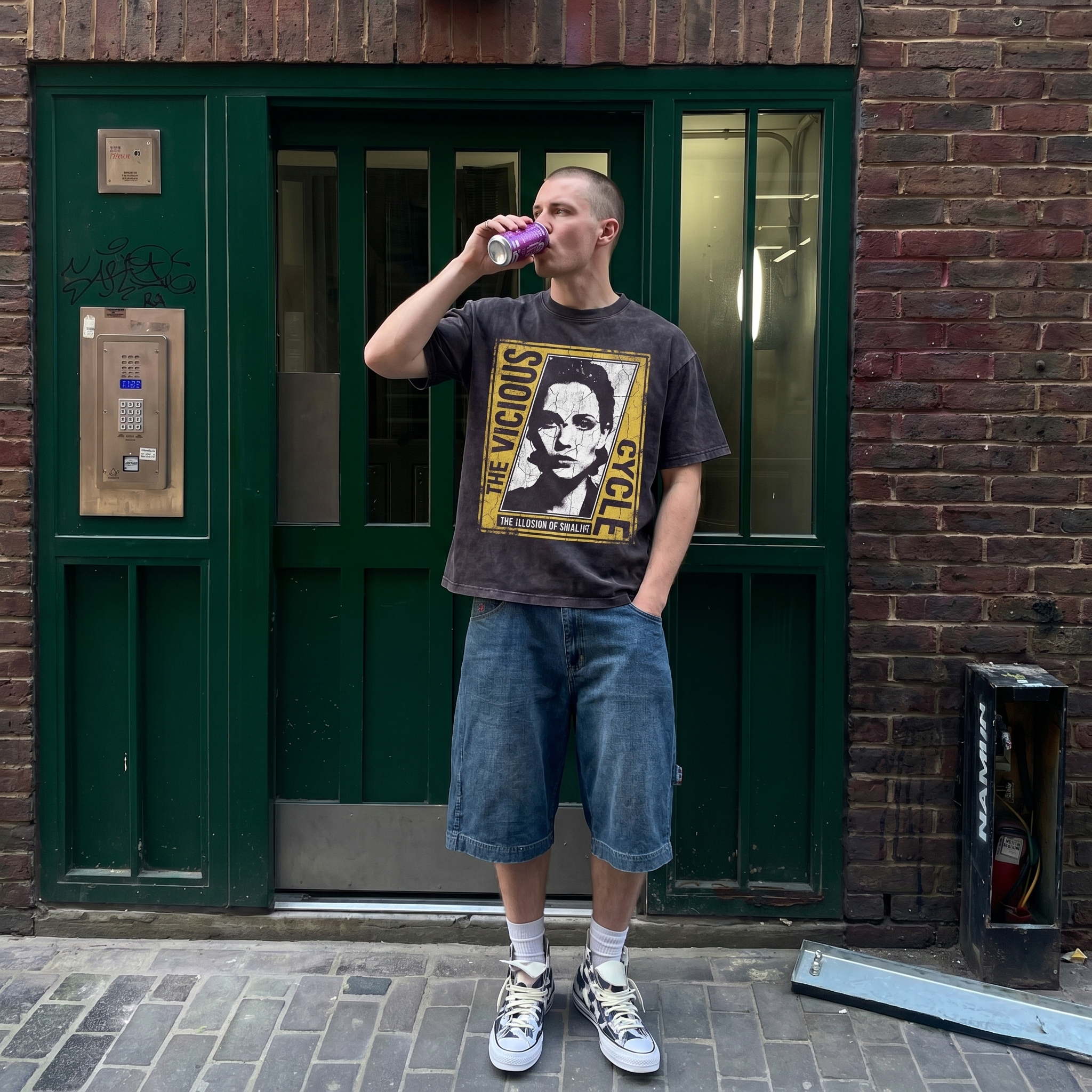 Man in graphic t-shirt and denim shorts drinks from can by green door in urban street